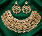An ornate gold-toned jewelry set arranged on a rich emerald green satin fabric background. The set includes a large choker necklace, matching drop earrings, and a round pendant with clear stones.